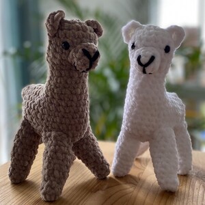 May include: Two handmade crochet llama figurines. One is a light brown color, the other is white. Both have black eyes and a black nose. The llamas are standing on a wooden surface.