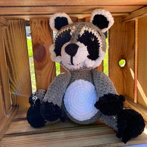 May include: A handmade crochet raccoon plush toy. The raccoon has a gray body, a white belly, and black accents around the eyes, ears, and paws. It is sitting inside a wooden crate.