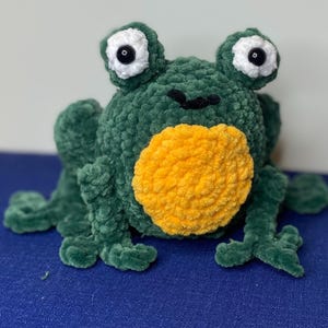 Op de afbeelding: Een groene pluche kikker met een gele buik en grote, witgerande ogen met zwarte pupillen. De kikker is gemaakt van een zacht, getextureerd materiaal en heeft vier poten. Een handgemaakt speelgoed.