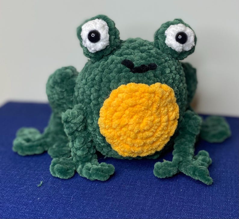 Op de afbeelding: Een groene pluche kikker met een gele buik en grote, witgerande ogen met zwarte pupillen. De kikker is gemaakt van een zacht, getextureerd materiaal en heeft vier poten. Een handgemaakt speelgoed.