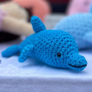 May include: A bright blue crocheted dolphin toy featuring a textured yarn surface, a single dorsal fin, side flippers, and a small black embroidered eye. The dolphin has a stitched mouth line and a rounded snout, crafted with visible yarn loops.