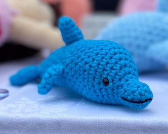 Op de afbeelding: Een felblauw gehaakt dolfijnspeeltje met een textuur van garen, een rugvin, zijvinnen en een klein zwart geborduurd oog. De dolfijn heeft een gestikte mondlijn en een afgeronde snuit, gemaakt met duidelijk zichtbare lussen van garen.