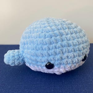 May include: A handmade, plush whale in light blue with white accents. The crocheted whale features small, black button eyes and a rounded body. The whale has a small tail and fins, perfect for a nursery or child's room.