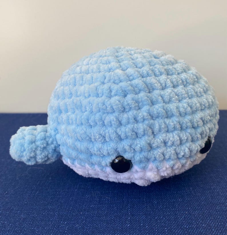 May include: A handmade, plush whale in light blue with white accents. The crocheted whale features small, black button eyes and a rounded body. The whale has a small tail and fins, perfect for a nursery or child's room.
