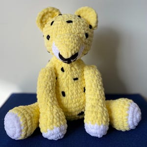 May include: A handmade, yellow crochet leopard plush toy with black spots. The toy has white accents on the muzzle and paws. The leopard is sitting with its legs extended and its ears perked up.