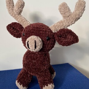 May include: A handmade plush moose toy in a sitting position. The moose is primarily a deep burgundy color with tan accents on the antlers, nose, and feet. The toy has small black eyes and a soft, textured appearance.