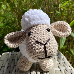 May include: A handmade crochet sheep toy with a fluffy white head and a tan body. The sheep has black eyes and a black stitched mouth. The toy is sitting on a woven surface, with green foliage in the background.