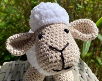 Op de afbeelding: Een handgemaakt gehaakt schaap speelgoed met een pluizige witte kop en een beige lichaam. Het schaap heeft zwarte ogen en een zwart gestikte mond. Het speelgoed staat op een geweven oppervlak, met groene bladeren op de achtergrond.