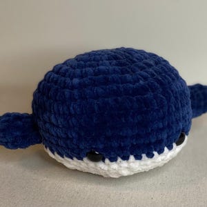 May include: A handmade, plush whale in a deep blue color. The whale has a rounded body, small fins, and a white belly. Black button eyes give the whale a friendly appearance. The whale is a soft, cuddly toy.
