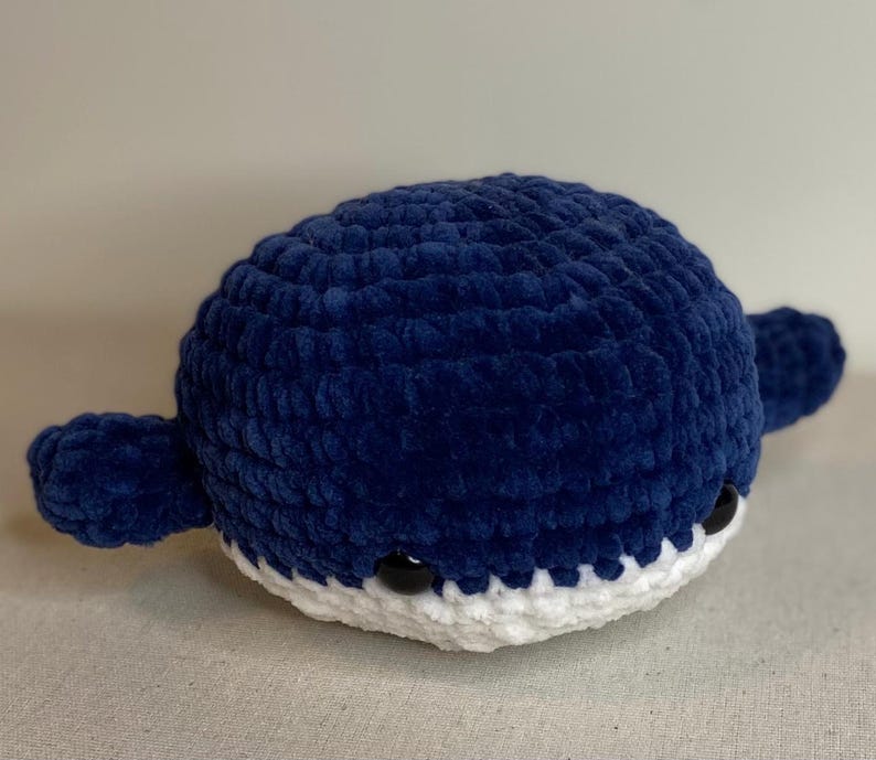 May include: A handmade, plush whale in a deep blue color. The whale has a rounded body, small fins, and a white belly. Black button eyes give the whale a friendly appearance. The whale is a soft, cuddly toy.
