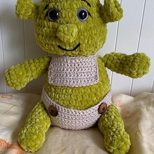 May include: A plush toy crafted from lime green yarn, featuring a rounded head with trumpet-shaped ears, large black eyes, and a stitched smile. The figure wears a light beige crocheted bib and matching diaper-style bottoms secured with two brown buttons.