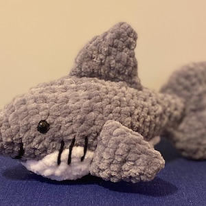 May include: A plush gray shark toy with a white belly and black accents. The shark has a dorsal fin, pectoral fins, and a tail fin. The toy is made of a soft, textured material and has black button eyes.