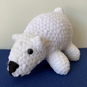 May include: A handmade white crochet polar bear with black eyes and a black nose. The plush toy is lying down on a blue surface. The bear has a textured, fluffy appearance.