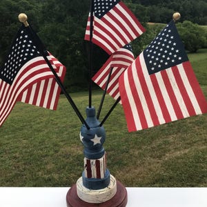 May include: A decorative wooden stand featuring a blue sphere with white stars and a red and white striped base, holding four small American flags on black poles with gold finials. The stand has a distressed finish with visible wood grain.