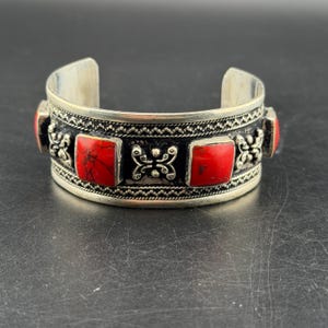 Pode incluir: Uma pulseira de punho em tom prateado com uma banda larga de padrões metálicos intrincados e quatro incrustações de pedras quadradas em vermelho brilhante. A superfície metálica apresenta um acabamento escuro e oxidado nas áreas rebaixadas, destacando os detalhes de volutas e as bordas em forma de corda que rodeiam as pedras quadradas vermelhas.