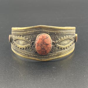 May include: A brass-toned cuff bracelet with an oval, reddish-orange stone at the center. The bracelet features intricate detailing, including twisted rope and floral patterns. Two smaller stones are set on the sides.