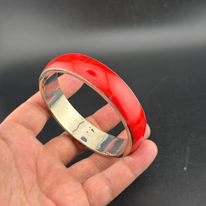 以下が含まれることがあります： 鮮やかな赤い象嵌細工が施された、銀色のバングルブレスレット。ブレスレットは滑らかで光沢のある仕上げで、大胆で人目を引くデザインです。赤い象嵌細工は、石または樹脂素材で作られているようです。