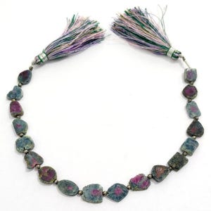 May include: A necklace featuring rough-cut gemstones in shades of blue and magenta. The stones are strung together with small silver beads. The necklace is finished with two tassels in shades of green, purple, and white.