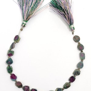 May include: A necklace featuring raw, multi-colored tourmaline beads in shades of green, pink, and gray. The necklace is accented with small silver-colored beads and finished with two decorative tassels with green, purple, and white threads.