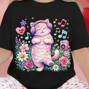 Puede incluir: Camiseta negra con un gato rosa con auriculares, rodeado de flores, notas musicales y un pequeño altavoz. El gato está de pie con los ojos cerrados, y el diseño tiene un estilo caprichoso y de dibujos animados.
