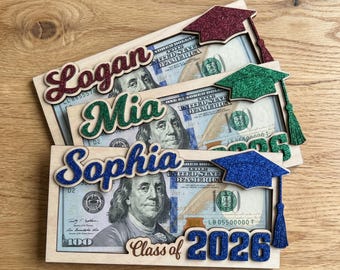 May include: Three wooden graduation gift holders, each with a $100 bill. The holders feature names in glittery script: Logan, Mia, and Sophia. Each has a glitter graduation cap and the text "Class of 2026".