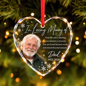 May include: A heart-shaped clear acrylic memorial ornament featuring a photo of an older man with a white beard. The text reads In Loving Memory of, Your life was a blessing, your memory a treasure. You are loved beyond words, and missed beyond measure, Dad, 1952-2026, with white snowflake accents and a red ribbon.