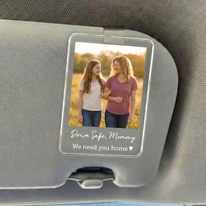 May include: A clear acrylic car visor photo holder with a picture of a woman and a girl. The photo holder has the text "Drive Safe, Mommy We need you home" in a script font.