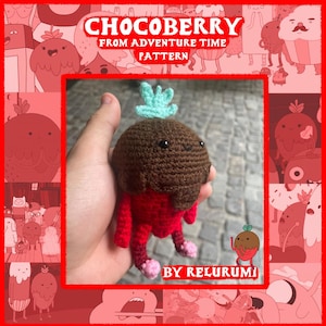 May include: A handmade crochet Chocoberry plushie from Adventure Time. The plushie features a brown head, red body, and light blue leaves. The text "CHOCOBERRY FROM ADVENTURE TIME PATTERN" is displayed in red. The plushie is held in a hand.