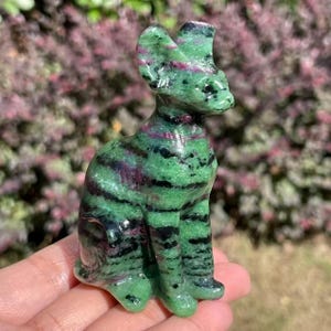 May include: A carved cat figurine made of green and black striped stone with red accents. The cat is sitting upright with its ears perked up. The figurine is held in a person's hand.