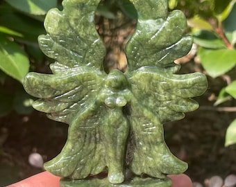May include: A green stone angel sculpture with outstretched wings. The figure has a detailed carved design, with visible feathers and a flowing gown. The sculpture is held in a hand, with a natural background of green foliage.