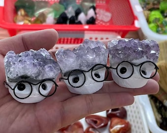 Puede incluir: Tres esculturas caprichosas de cristal de amatista, cada una con una base blanca, gafas con montura negra y ojos grandes negros. Los cristales de amatista son una mezcla de púrpura y transparente. Las esculturas se sostienen en una mano.