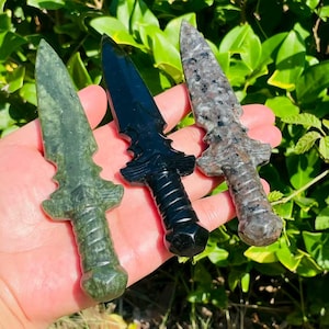 May include: Three carved stone daggers in shades of green, black, and gray. Each dagger features a detailed handle and a pointed blade. The daggers are displayed against a backdrop of green foliage.