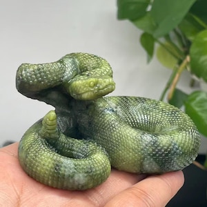 Puede incluir: Una escultura detallada de una serpiente enroscada en tonos verdes. Las escamas de la serpiente están intrincadamente talladas, y su cabeza está levantada, mostrando sus ojos y boca. La escultura está hecha de un material similar a la piedra.