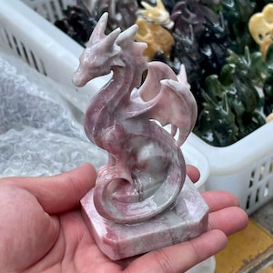 Peut inclure: Une figurine de dragon rose et blanc sculptée. Le dragon a des ailes détaillées, un long cou et est perché sur une base carrée. La figurine est tenue dans la main d'une personne, mettant en valeur les détails complexes de la sculpture.