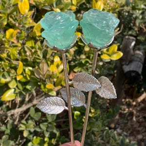 May include: Two decorative rose stems with light teal-colored rose blooms. The stems are gold-colored with silver-colored leaves. The background is a blurred view of green and yellow foliage.