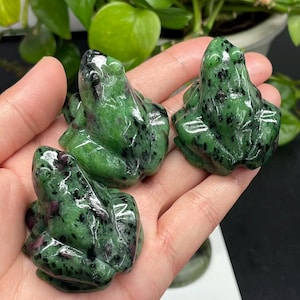 Op de afbeelding: Drie gepolijste kikkerbeeldjes. De kikkers zijn groen met zwarte en rode spikkels. De beeldjes worden in een hand gehouden, met een vage achtergrond van groen gebladerte.