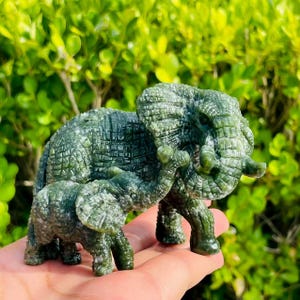 Puede incluir: Una figura de elefante de piedra verde tallada con una cría de elefante más pequeña. El elefante adulto tiene detalles, incluyendo una superficie texturizada y colmillos visibles. La cría está colocada cerca del adulto, sugiriendo un tema familiar.
