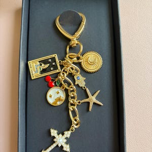 K&ouml;nnte beinhalten: Goldfarbener Schl&uuml;sselanh&auml;nger mit verschiedenen Anh&auml;ngern, darunter ein Kreuz, ein Seestern, ein Sonnenstrahl und eine Tarotkarte. Der Schl&uuml;sselanh&auml;nger befindet sich in einer schwarzen Box mit einem goldenen Verschluss und einer Kette. Die Anh&auml;nger sind mit Emaille und Kristallen verziert.