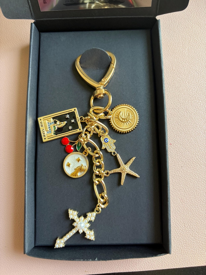 K&ouml;nnte beinhalten: Goldfarbener Schl&uuml;sselanh&auml;nger mit verschiedenen Anh&auml;ngern, darunter ein Kreuz, ein Seestern, ein Sonnenstrahl und eine Tarotkarte. Der Schl&uuml;sselanh&auml;nger befindet sich in einer schwarzen Box mit einem goldenen Verschluss und einer Kette. Die Anh&auml;nger sind mit Emaille und Kristallen verziert.