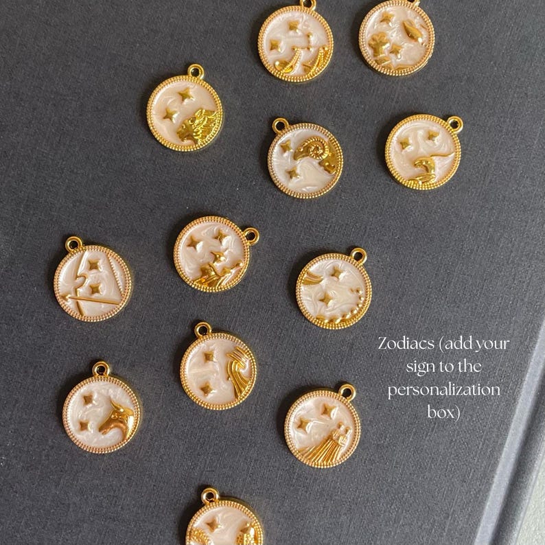 K&ouml;nnte beinhalten: Goldfarbene Tierkreiszeichen-Anh&auml;nger mit wei&szlig;en und goldenen Akzenten. Jeder Anh&auml;nger zeigt ein anderes Tierkreiszeichen und Sterne. Die Anh&auml;nger sind rund mit einer kleinen Schlaufe oben.