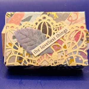 May include: A small, decorative box with a floral patterned design. The box is adorned with a gold filigree overlay and a light blue fabric leaf. The text "the loveliest things" is displayed on a white label.