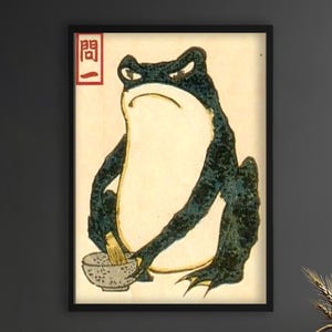Könnte beinhalten: Gerahmter Kunstdruck mit einer grimmigen Froschdarstellung im traditionellen japanischen Stil. Der Frosch ist dunkelgrün und schwarz und hält eine Schüssel. Das Kunstwerk hat eine Vintage-Ästhetik mit cremefarbenem Hintergrund und schwarzem Rahmen.