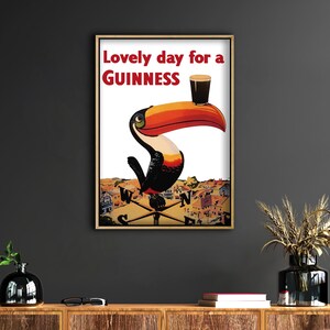 May include: Framed vintage-style poster featuring a toucan with a Guinness pint on its beak. The poster has the text "Lovely day for a Guinness" in red. The toucan is perched on a weather vane above a town.