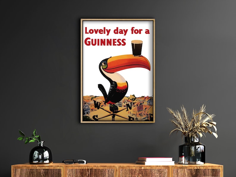 May include: Framed vintage-style poster featuring a toucan with a Guinness pint on its beak. The poster has the text "Lovely day for a Guinness" in red. The toucan is perched on a weather vane above a town.