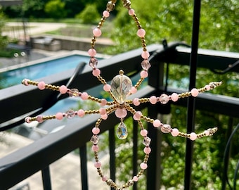 May include: A decorative suncatcher shaped like a spider, made with pink beads, clear crystals, and gold-tone wire. The spider has eight legs and a central crystal body. It is designed to catch the sunlight and create a sparkling effect.