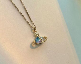 May include: Gold-toned necklace featuring a pendant shaped like a planet with a blue and purple center and a ring of small crystals. The chain is delicate and the pendant is small, creating an elegant and celestial-themed accessory.