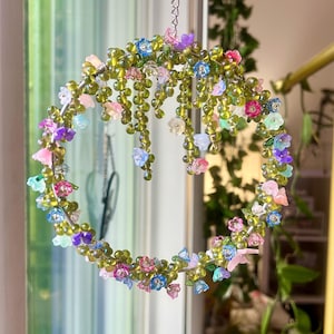 May include: A beaded floral wreath with a chain for hanging. The wreath is adorned with small, colorful flowers in shades of pink, blue, purple, and green. The beads are olive green and the flowers are made of plastic.