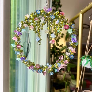 May include: A beaded floral wreath with a silver chain for hanging. The wreath is adorned with colorful, translucent flowers in shades of pink, blue, purple, and green. The wreath is suspended in front of a window.