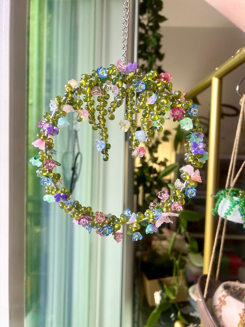 May include: A beaded floral wreath with a silver chain for hanging. The wreath is adorned with colorful, translucent flowers in shades of pink, blue, purple, and green. The wreath is suspended in front of a window.