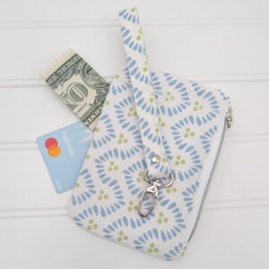 Puede incluir: Un monedero con estampado blanco y azul con cremallera plateada y correa de muñeca. Un billete de un dólar y una tarjeta de crédito son parcialmente visibles en el interior. El monedero tiene un cierre plateado.